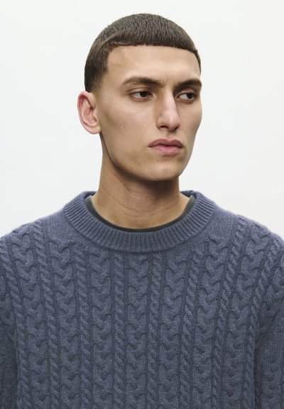 Tmavě modrý svetr s pletenými vzory a vroubkovaným kulatým výstřihem. Texturovaný vzor obsahuje vyvýšené copánky, což zdůrazňuje silný a pohodlný design.