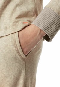 Beige cotton long-sleeve top with ribbed cuffs and a small orange label near the hem; hand in pocket shows relaxed fit design.