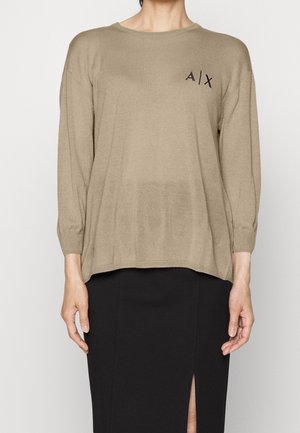 Person iført beige langærmet sweater med "A|X" logo på brystet og sort nederdel med frontslids, stående mod en ensfarvet baggrund.