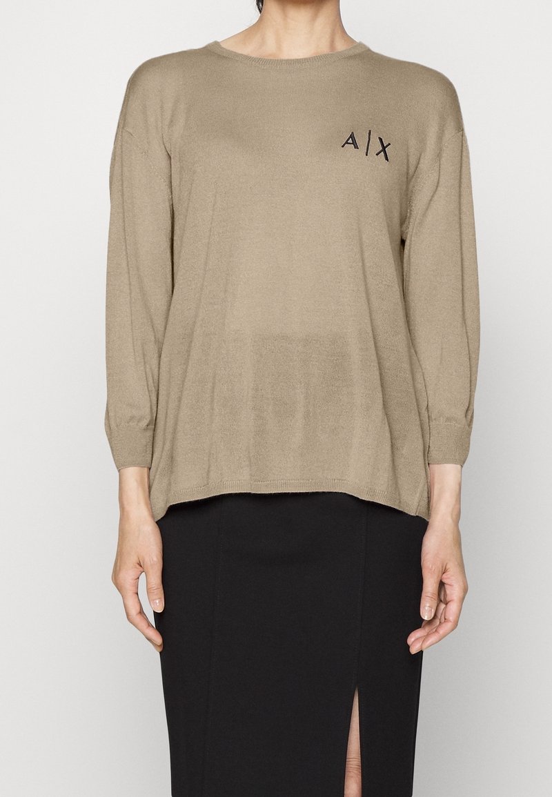 Personne portant un pull à manches longues beige avec le logo "A|X" sur la poitrine et une jupe noire avec une fente à l'avant, debout devant un fond uni.