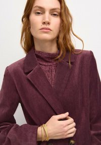 Plum herringbone bleiseri suurte klappidega ja kahe nupuga, kihtide kaupa tekstuuriga ploomivärvi kõrge kaelusega pluusi peale. Kuldsed käevõrud kaunistavad randmeid.