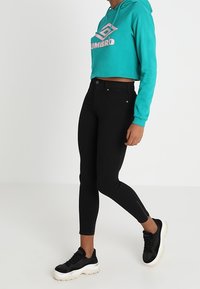 Sudadera corta turquesa con logo rosa, combinada con jeans skinny negros. Zapatillas negras con suelas blancas. Textura de tela suave y diseño ajustado.