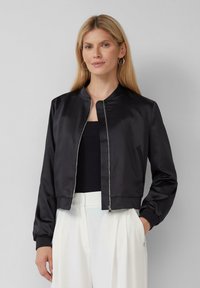 Giacca bomber nera in satin con zip frontale, maniche a costine e design cropped. Indossata sopra una maglietta nera, abbinata a pantaloni bianchi.