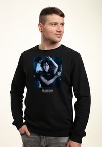 Man med svart sweatshirt med en grafisk bild av en kvinna i en svart klänning som dansar, texten "Onsdag" under bilden.