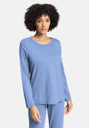 Donna con capelli ricci che indossa una maglia a maniche lunghe azzurro chiaro e pantaloni abbinati, in piedi davanti a uno sfondo bianco semplice.
