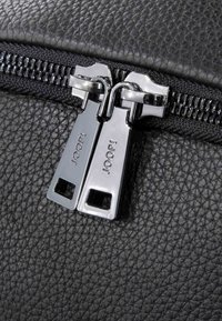 Černá kožená taška s texturou obsahující dvojité stříbrné zipy s gravírovaným logem na táhlech, podtrhující robustní a stylový design uzávěru.