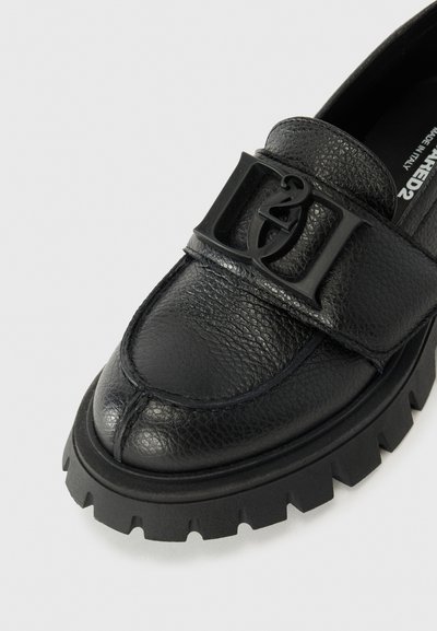 Svart läder loafer med texturerad yta, som har en uttalad logobuckla, rund tå och kraftig svart gummisula med ribbade kanter.