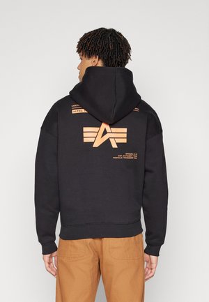 Mann med korte dreadlocks iført en svart hettegenser med oransje logo og tekst, kombinert med lysebrune bukser, sett bakfra.