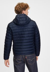 Marinblå puffjacka med huva, med quiltade sektioner och en slät textur, bärs över mörka jeans med synliga söm detaljer.