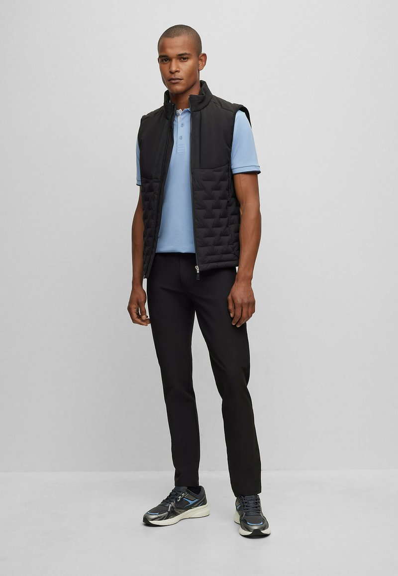 Gilet rembourré noir avec un col haut sur un polo bleu clair. Pantalon noir slim-fit et baskets grises avec des accents réfléchissants.