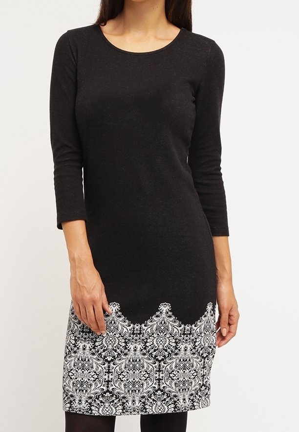 Femme portant une robe noire longueur genoux avec des manches trois quarts et un motif floral blanc le long de l'ourlet.
