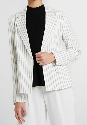 Person iført en hvid blazer med tynde vertikale grå striber over en sort top og hvide bukser, der holder blazers revers med den ene hånd.