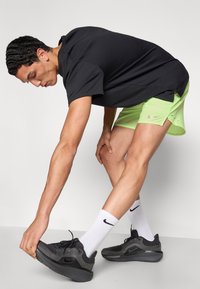 Μαύρο αθλητικό μπλουζάκι, νέον πράσινα σορτς και μαύρα αθλητικά παπούτσια Nike με γκρι σόλα, σε πλάγια όψη καθώς το άτομο τεντώνεται.