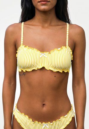 Mujer que lleva un bikini a rayas amarillas y blancas con bordes ondulados y pequeños lazos blancos en la parte superior e inferior.