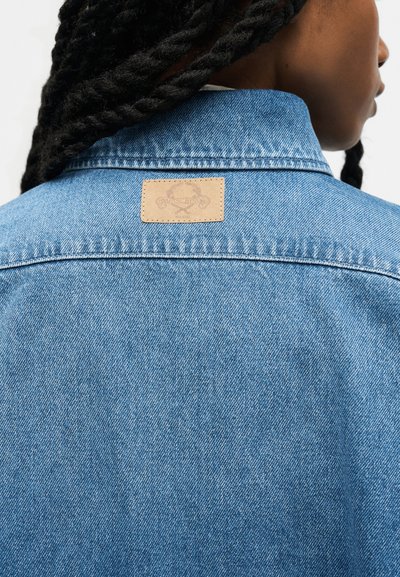 Personne aux longs cheveux tressés portant une veste en jean bleu clair montrant une petite étiquette en cuir beige sur le haut du dos.