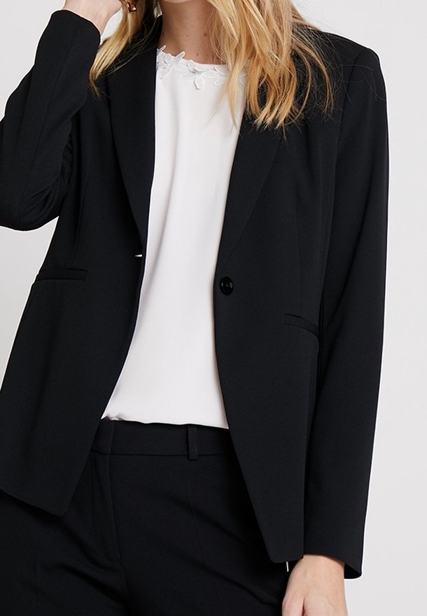 Femme portant un blazer noir et un pantalon assorti sur un chemisier blanc avec un détail floral au niveau du décolleté, debout devant un fond uni.