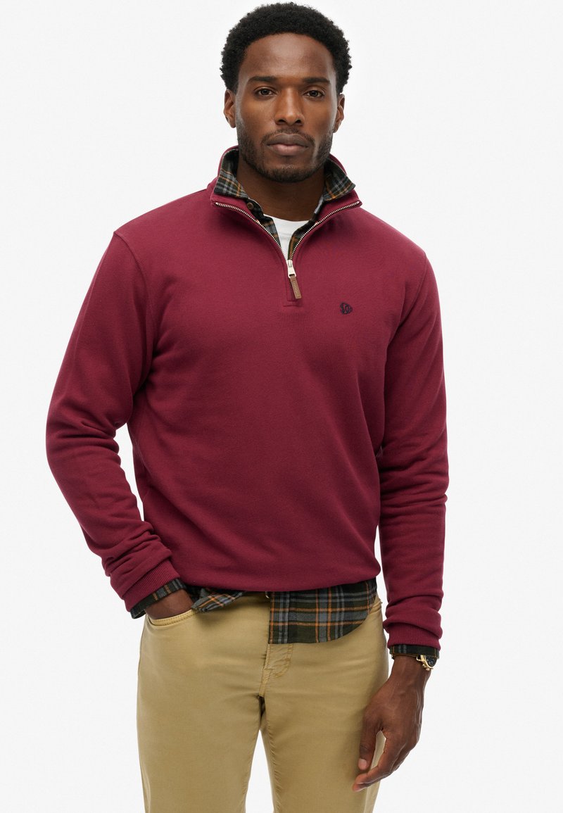 Bordowy sweatshirt z krótkim zamkiem i miękką fakturą, z małym logo na piersi, noszony na koszuli w kratę.