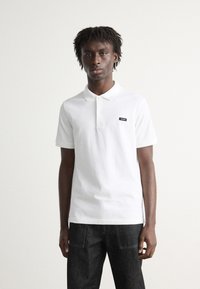 Weißes Polohemd aus Baumwolle mit kragen, kurzen Ärmeln und einem kleinen schwarzen Logo-Patch auf der linken Brust. Dunkle Jeans runden den Look ab.