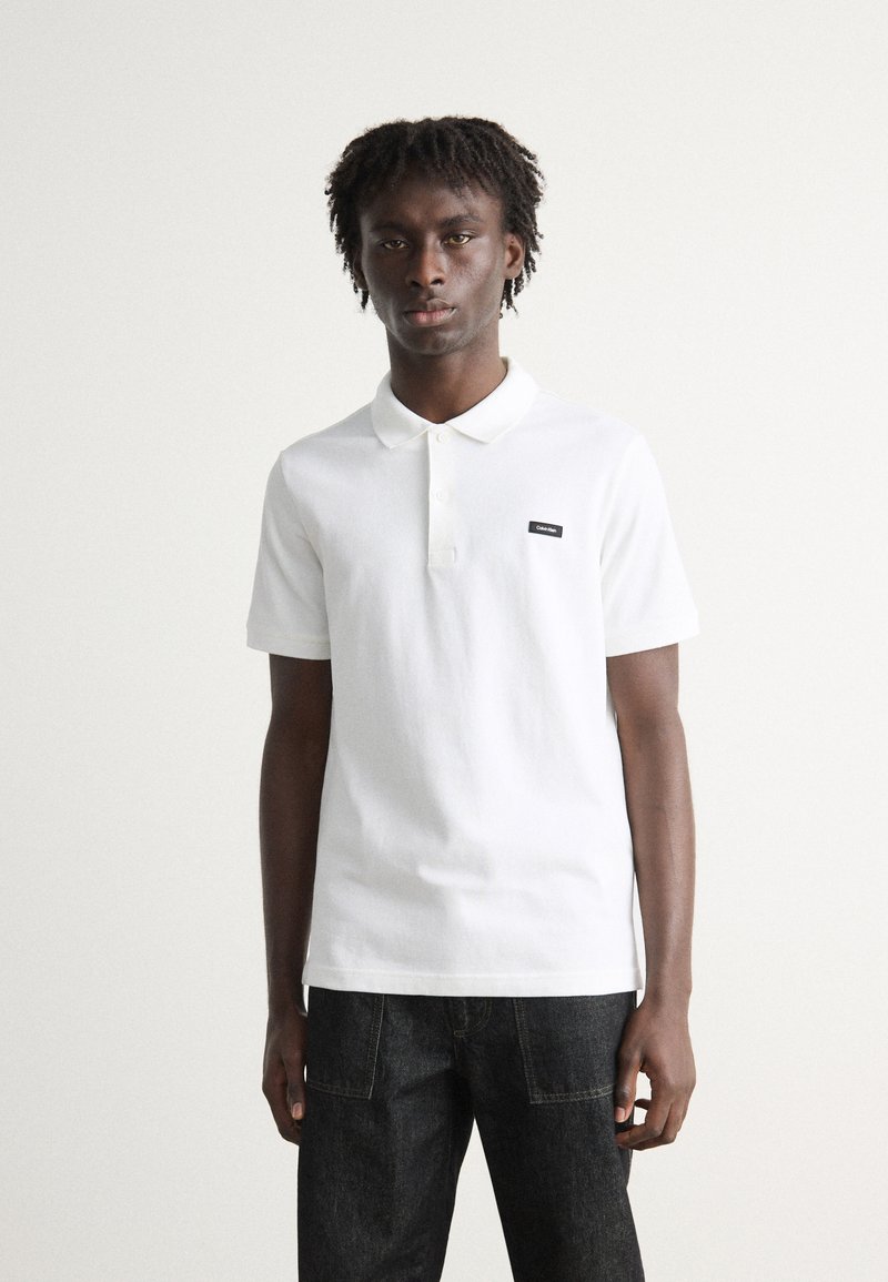 Weißes Polohemd aus Baumwolle mit kragen, kurzen Ärmeln und einem kleinen schwarzen Logo-Patch auf der linken Brust. Dunkle Jeans runden den Look ab.