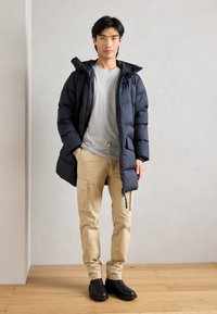 Tengerészkék pufikabát kapucnival, szürke póló, bézs cargo nadrág és fekete bokacsizma. Cipzáras zsebekkel és steppelt textúrával rendelkezik.