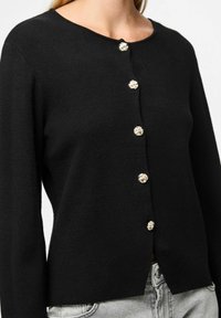 Melnā adīta kardigāna, kuram ir apaļa apkakle un piecas dekoratīvas zelta krāsas pogas ar ziedu rotājumiem priekšpusē.