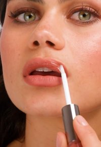 Donna con occhi verdi che applica un lucidalabbra nude al labbro inferiore con un applicatore bianco, primo piano sul viso e sulle labbra.