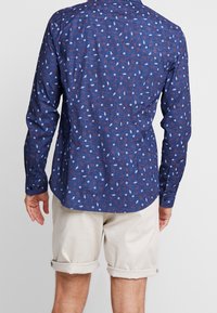 Koszula z długim rękawem w ciemnoniebieskim kolorze z wzorem liści w białym i czerwonym. Teksturowany materiał z zapięciem na guziki przy mankietach i zaokrąglonym dołem.