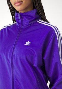 Donna che indossa una giacca da ginnastica Adidas viola con strisce bianche sulle maniche e un logo Adidas bianco sul petto, chiusa fino al collo.