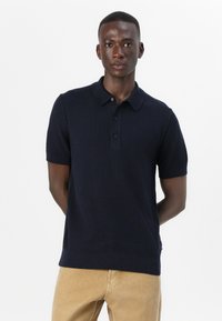 Polo shirt bleumarin cu tricot texturat, mâneci scurte și o pată cu trei nasturi. Asortat cu pantaloni bej.