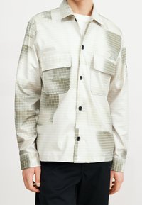 Camisa de cuello en beige claro con un patrón de cuadrícula y acentos olivo. Presenta dos bolsillos en el pecho y botones negros. Textura suave y elegante.