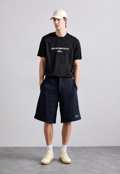 Juodas medvilninis marškinėliai su "Emporio Armani Milano" logotipu, derinami su tamsiai mėlynomis šortais ir kremiškai baltomis sportiniais batais. Paprasta kepurė užbaigia aprangą.