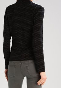 Černý top s dlouhým rukávem a přiléhavým střihem, zdobený malými barevnými puntíky. Látka se zdá být hladká s mírným leskem.