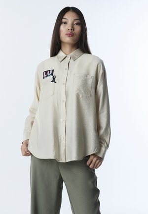 Junge Frau mit langen dunklen Haaren, die ein beigefarbenes Hemd mit Knöpfen und einem "LUCK"-Aufnäher sowie dunkle Hosen trägt, vor einem schlichten weißen Hintergrund steht.