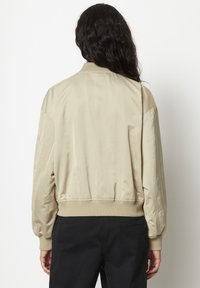Eine Frau mit langen, dunklen Haaren steht mit dem Rücken zur Kamera, trägt eine beige Bomberjacke und schwarze Hosen vor einem schlichten weißen Hintergrund.