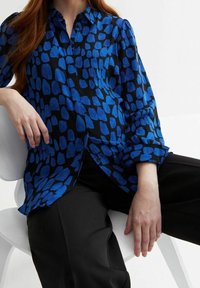 Blue and black blouse featuring a spotted pattern, long sleeves, and a collared neckline. Smooth fabric and relaxed fit.