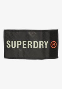 Czarna tkaninowa opaska na nadgarstek z napisem "SUPERDRY" bold białymi literami oraz czerwonym znakiem zastrzeżonym po prawej stronie.