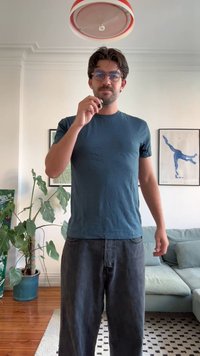 Tyrkysové bavlněné tričko s krátkým rukávem, spárované s tmavě šedými džíny. Postoj ve vzpřímené poloze, drží nějaký předmět, vnitřní prostředí s zelenými rostlinami.