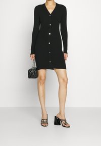 Robe noire à manches longues côtelée avec un col en V et des boutons sur le devant, associée à des sandales à talons noires à brides et un petit sac noir.