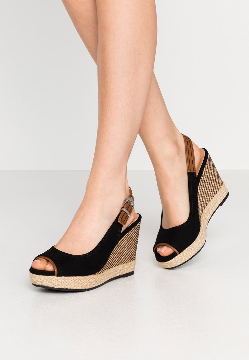 Jambes portant des sandales compensées noires et marron avec bouts ouverts et brides arrière sur un sol gris clair.