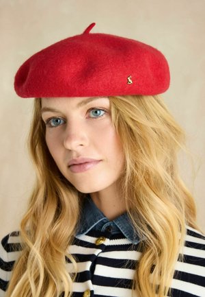 Rotes Woll-Baskenmütze mit kleinem oberem Schlaufe und goldener Anstecknadel. Das Modell trägt ein gestreiftes Hemd mit Jeanskragen. Weiche Textur.