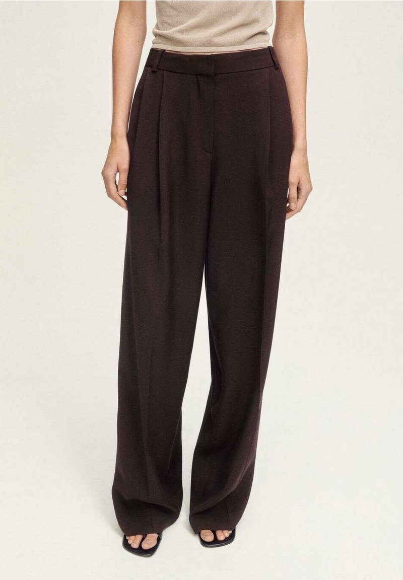 Pantalon marron foncé taille haute à jambes larges porté avec un haut beige sans manches et des sandales noires à bouts ouverts sur un fond uni.