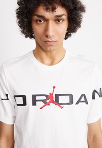 Tricou alb din bumbac, cu un mare imprimeu negru "JORDAN" și un logo Jumpman roșu pe față; guler rotund și mâneci scurte.