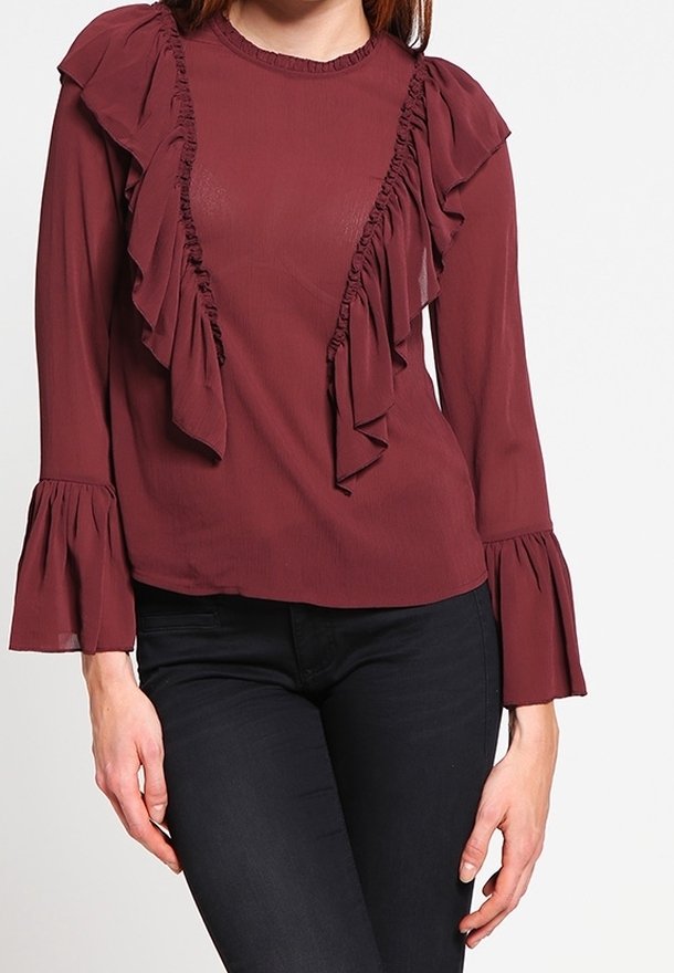Femme portant un chemisier bordeaux à manches longues avec des détails à volants sur le devant et des manches évasées, associé à un pantalon noir.