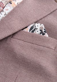 Struktureret mauve blazer med en revers med hak og et blomsterprintet lommetørklæde med sorte og taupe mønstre. Synlige syningsdetaljer.
