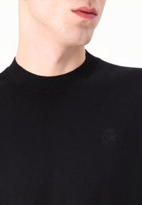 Svart strikkegenser med rund hals, med en subtil logo på brystet og en myk tekstur.