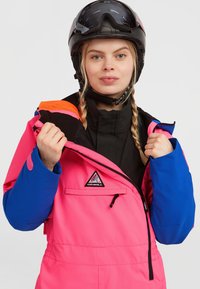 Heldere roze skijas met blauwe accenten, rits voorzak en logo-opdruk. Het materiaal lijkt waterdicht en geïsoleerd. Draagt met een zwarte helm.