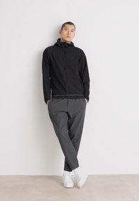 Črna zip-up jakna s kapuco, v kombinaciji s sivimi ožjimi hlačami in belimi supergami. Gladek material z minimalističnim dizajnom.