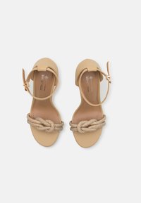 Beige vegansk sandal med knyttet, perlebesatt rem og ankelspenne. Glatt overmateriale, åpen tådesign, flat såle.