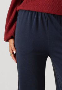 Pantaloni con elastico in vita di colore blu navy, dalla texture liscia, silhouette aderente e dettagli con spacco laterale. Mano appoggiata sulla coscia.