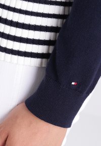 Granatowy sweter z poziomymi białymi paskami, prążkowana faktura i małe logo w kolorach czerwonym, białym i niebieskim na mankiecie.